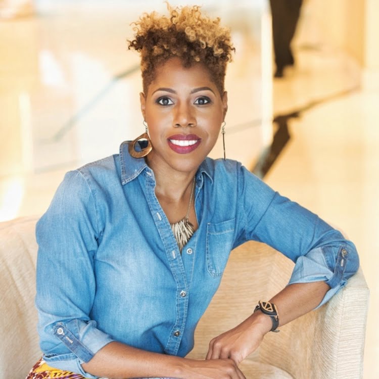 Andrene M. Taylor with short curly hair wearing a denim shirt and gold jewelry sits on a beige chair, looking at the camera with a slight smile—embodying the spirit to decolonize African storytelling through her confident presence. - Kipaji Lab