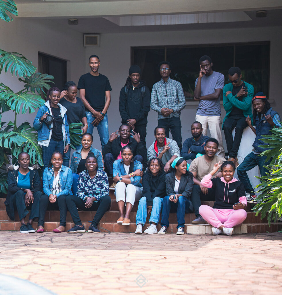 A group of nineteen people pose for a photo on outdoor steps in front of a building, some standing, some sitting, with plants on either side—gathered for creative training in Kenya. - Kipaji Lab