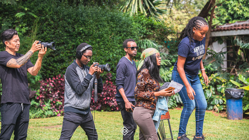 Five people are outdoors in lush greenery; two hold cameras, one has papers, and another stands on a chair while others watch—a scene alive with creative training Kenya style. - Kipaji Lab