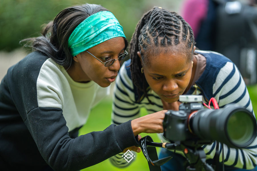 Two women closely examining and adjusting the settings on a professional camera outdoors during a creative training Kenya session. - Kipaji Lab