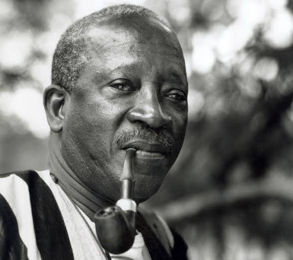 Black and white photo of an older man in a striped shirt, holding a pipe in his mouth, looking slightly away from the camera at the Red Balloon Festival Africa. - Kipaji Lab