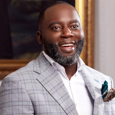 A man with a short beard wearing a light plaid suit jacket, white shirt, and pocket square smiles at the camera, embodying the spirit to decolonize African storytelling through style and confidence. - Kipaji Lab